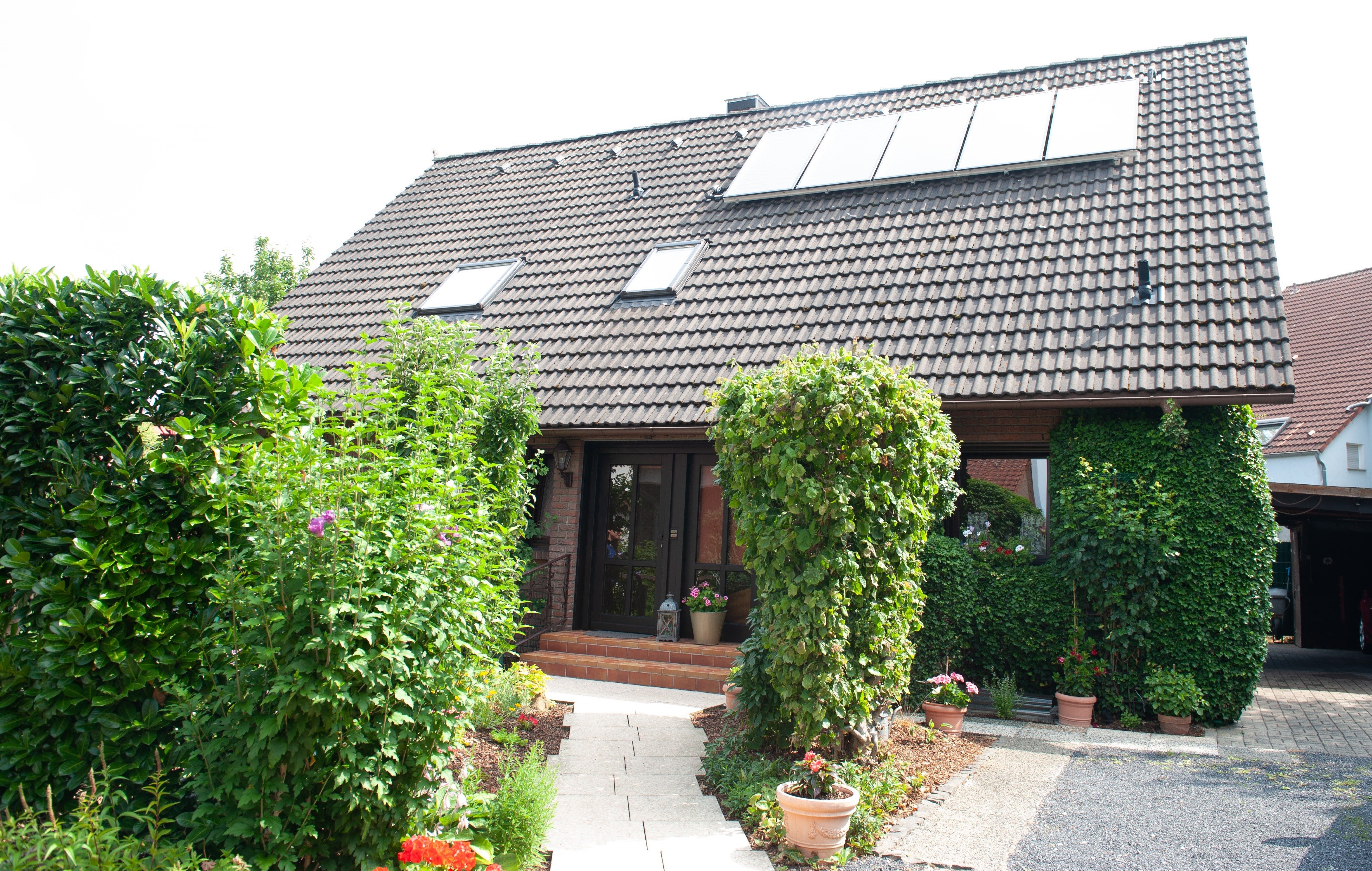 Dieses Bild zeigt ein Bild von einem Wohnhaus mit Solarplatten zu sehen. Davor ist ein Garten und im Hintergrund ein weiteres Wohnhaus zu sehen.