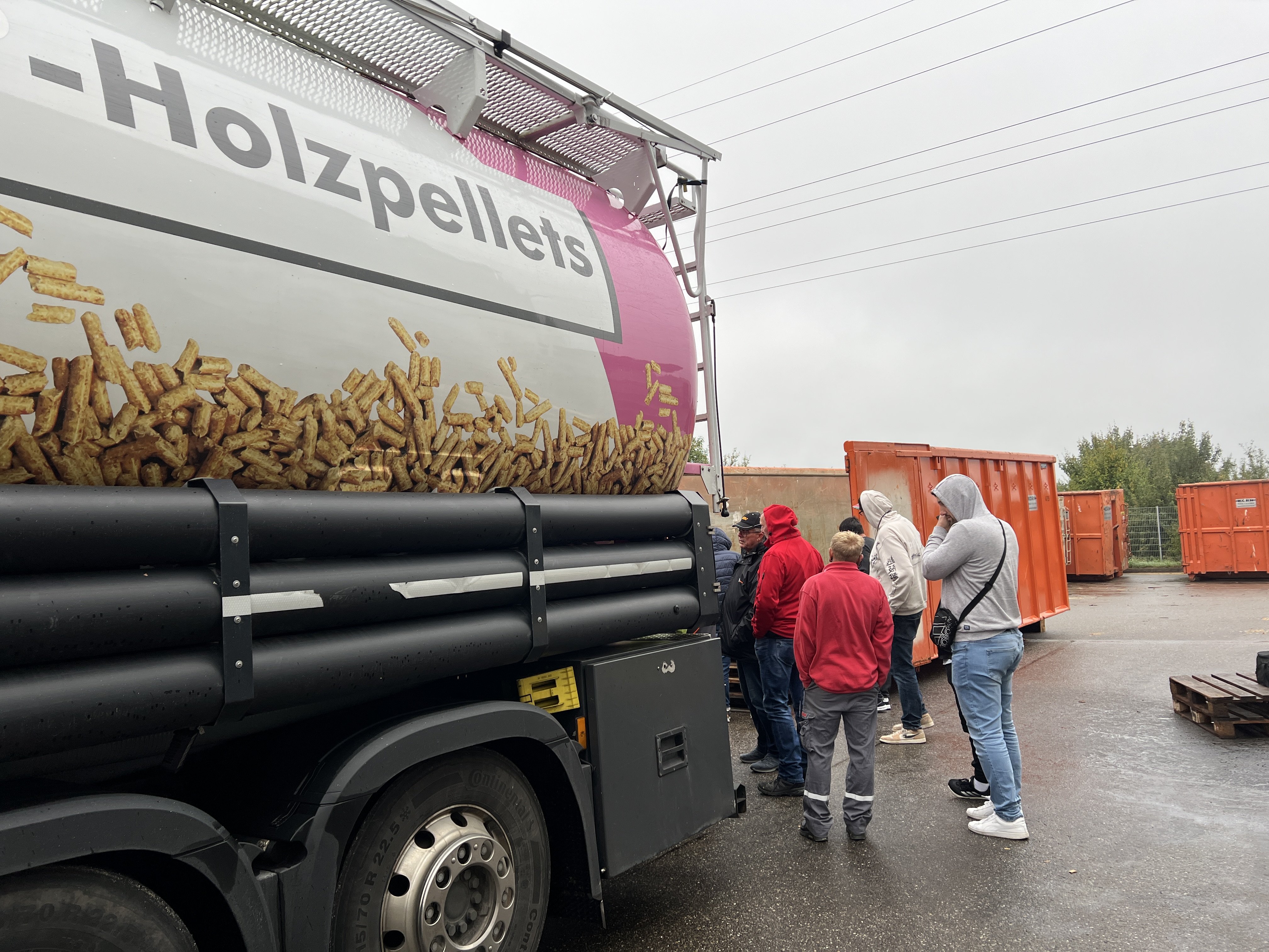 Das Bild im Querformat zeigt einen pinken Lkw mit Pellets. Daneben eine Gruppe Menschen im Gespräch.
