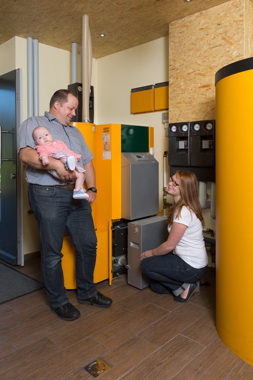 Das Bild zeigt eine Familie mit Kleinkind im Kellerraum vor einem gelben Pelletkessel.