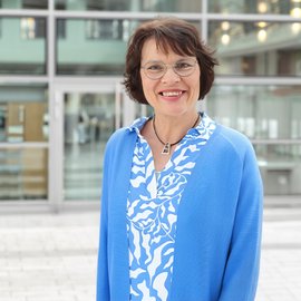 Frau steht vor einem Gebäude. Sie trägt eine leuchtend blaue Jacke.