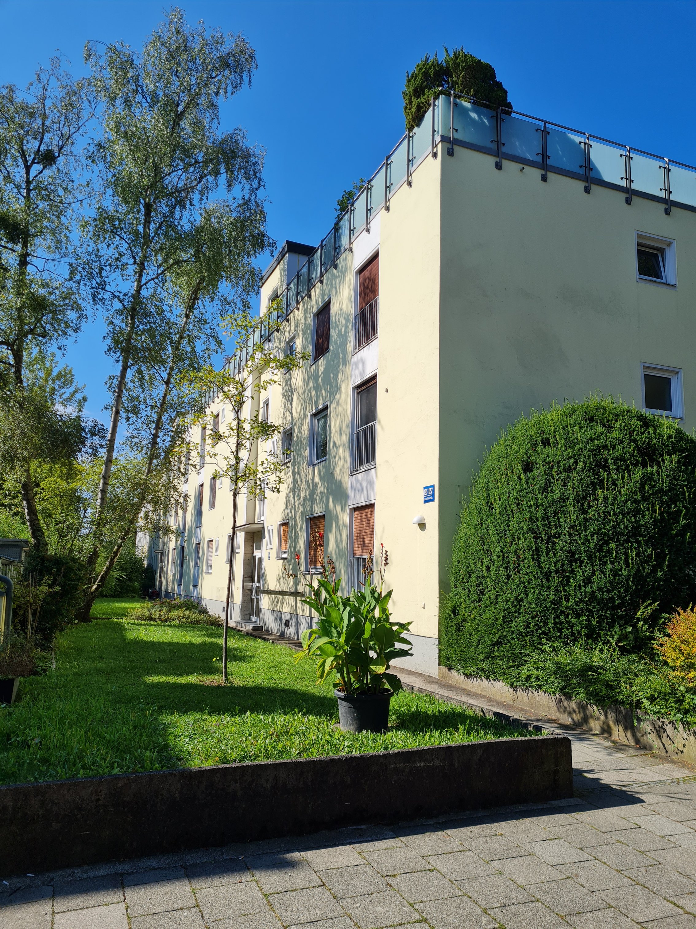 Das Bild zeigt ein gelbes Wohnhaus (rechts). Im Vordergrund ist ein Vorgarten mit Gehweg und im Hintergrund ein blauer Himmel zu sehen.
