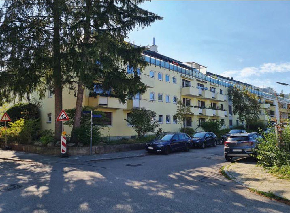 Das Bild zeigt ein Text von einem gelben Wohnhaus. Davor ist eine Straße und im Hintergrund ein blauer Himmel zu sehen.