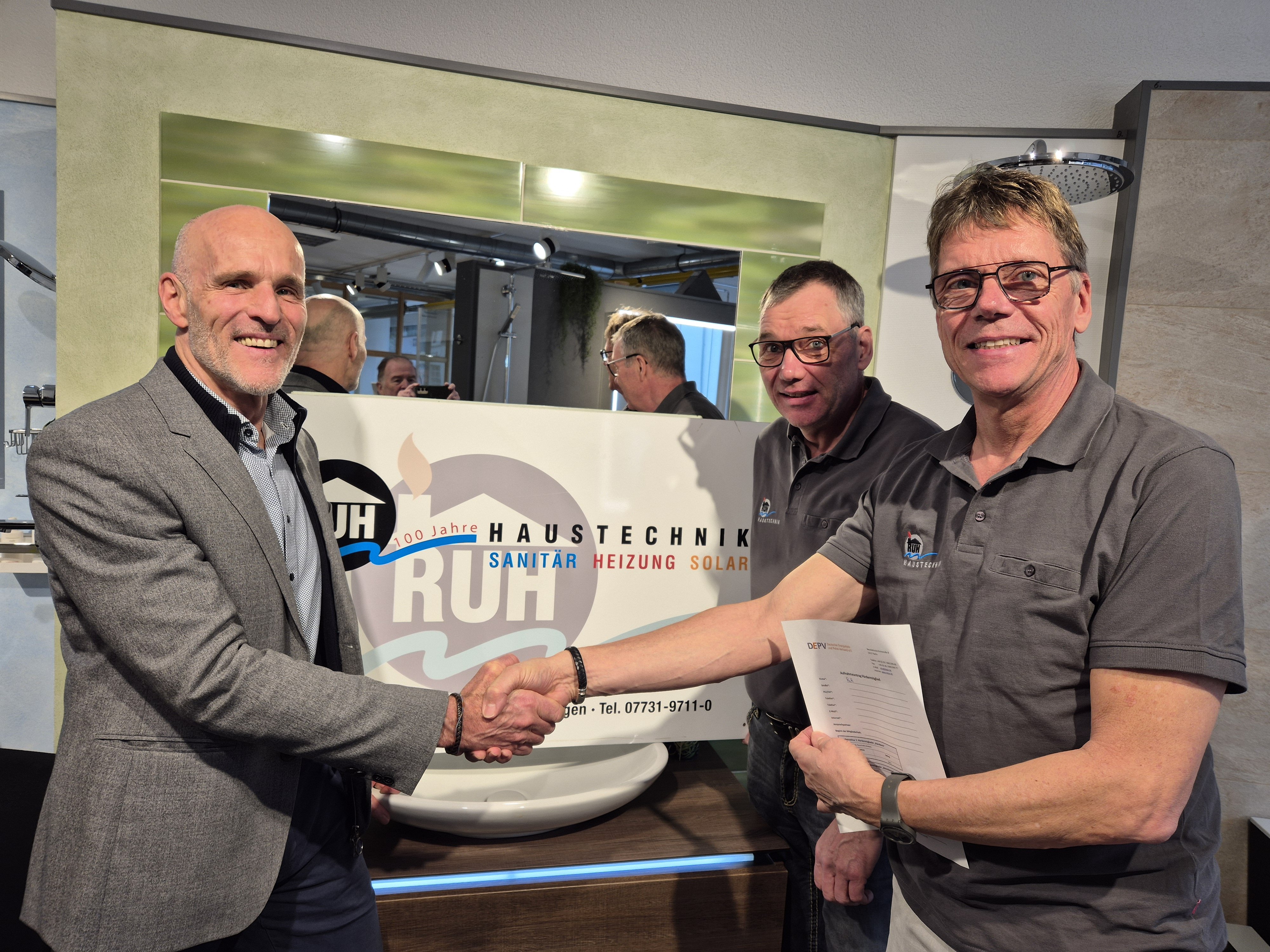 Drei Männer, zwei im Vordergrund, einer rechts im iIntergrund, die beiden im Vordergrund schütteln sich die Hand vor einem Firmenschild