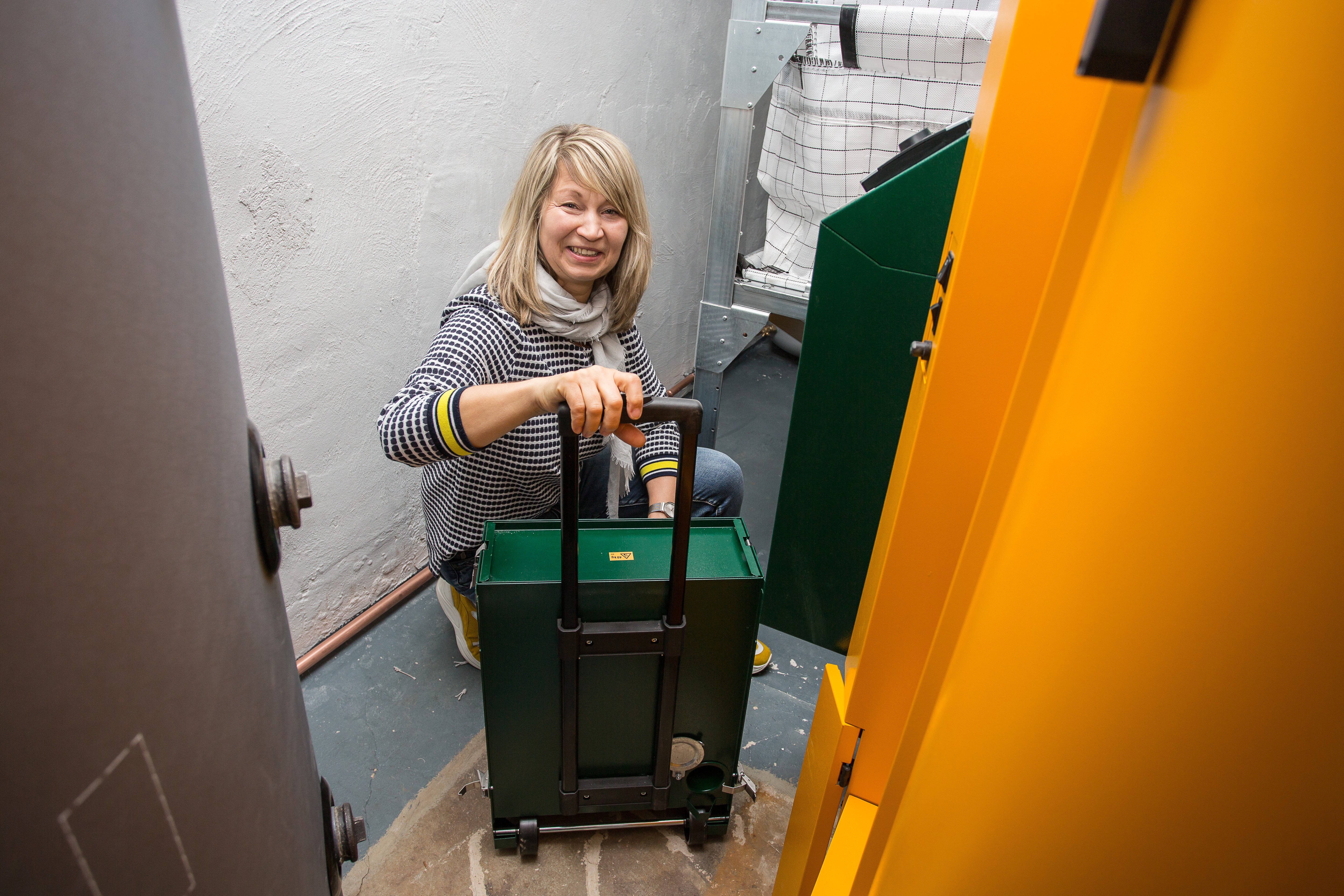 Das Bild zeigt eine weibliche Person mit einem Aschetrolley im Kellerraum vor einem grünen Pelletkessel.